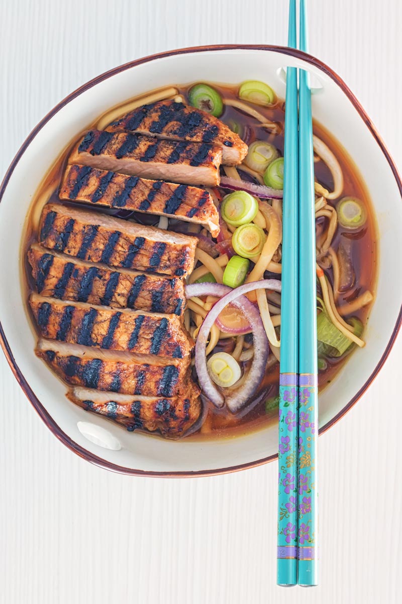 Overhead portrait image of a spicy pork ramen noodle soup served in a bowl with turquoise chopsticks