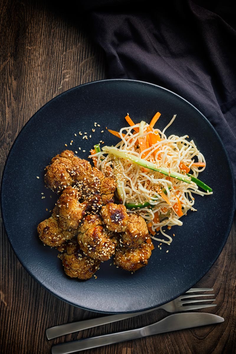 Tall overhead image of spicy roasted cauliflower florets with a noodle salad side dish on a black plate