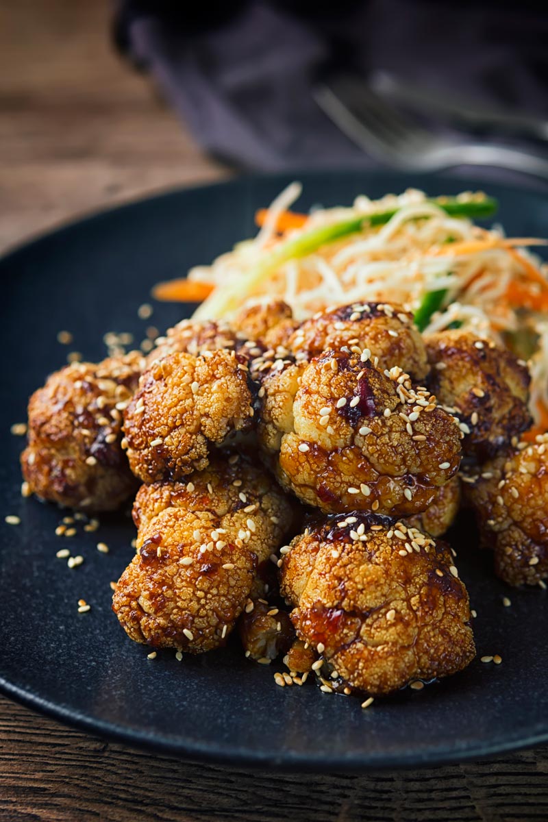 Tall image of spicy roasted cauliflower florets with a noodle salad side dish on a black plate