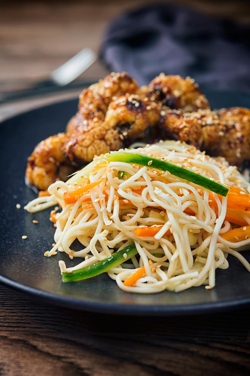 Tall image of a noodle salad served with roasted cauliflower florets on a black plate