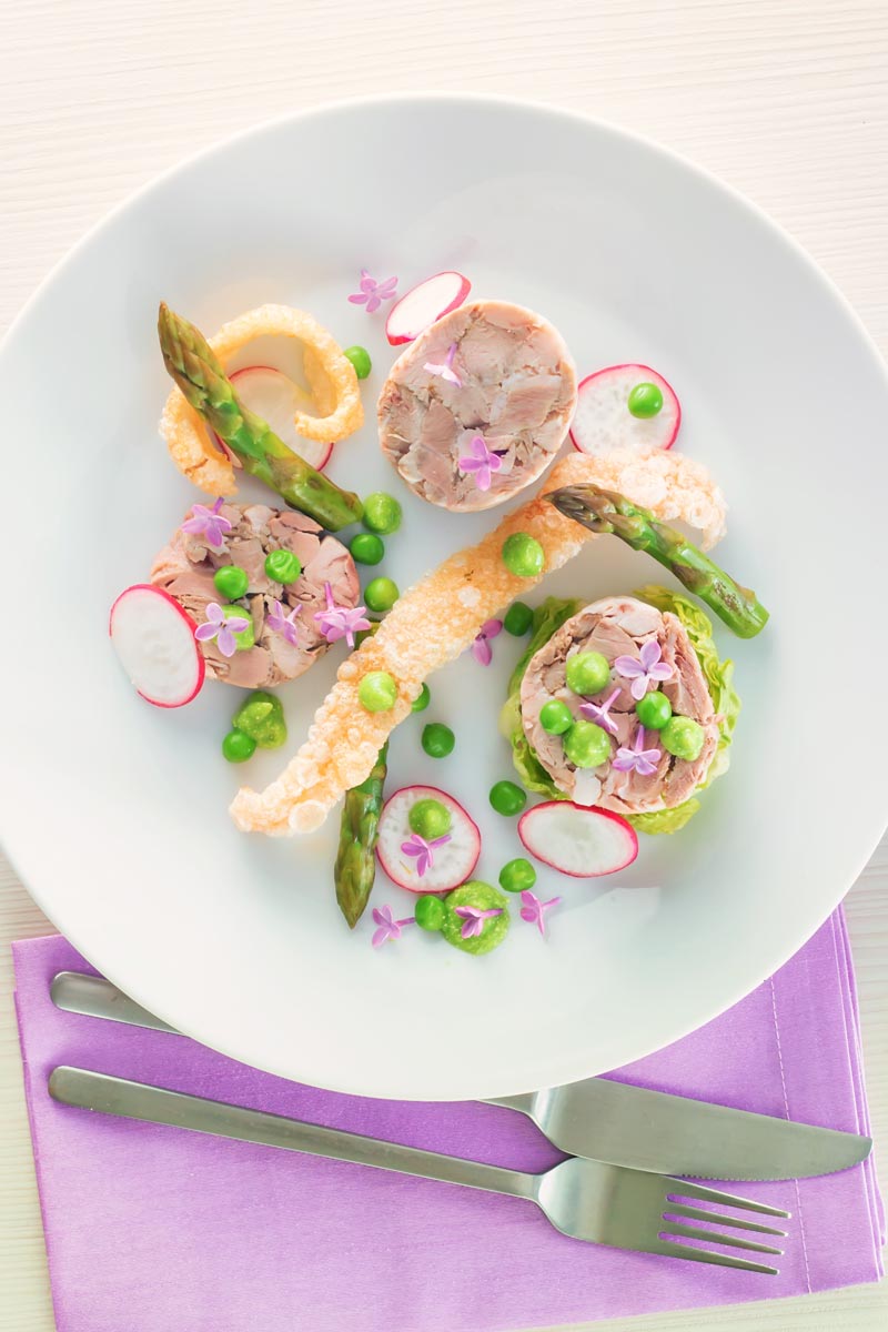 Portrait overhead image of a spring ham hock salad, featuring pressed ham hock, asparagus, peas, lettuce, radish, and puffed pork crackling.