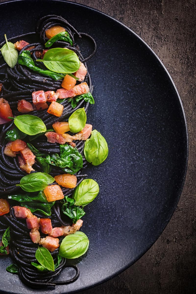 Tall over head image of black squid ink pasta with bacon, spinach and basil plated elegantly on a black plate against a dark backdrop