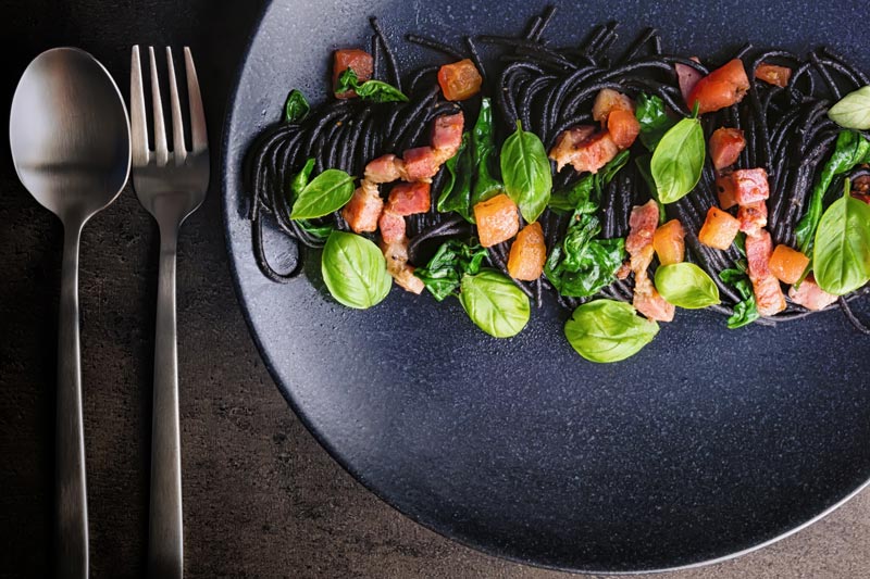 Landscape over head image of black squid ink pasta with bacon, spinach and basil plated elegantly on a black plate against a dark backdrop with a fork and spoon