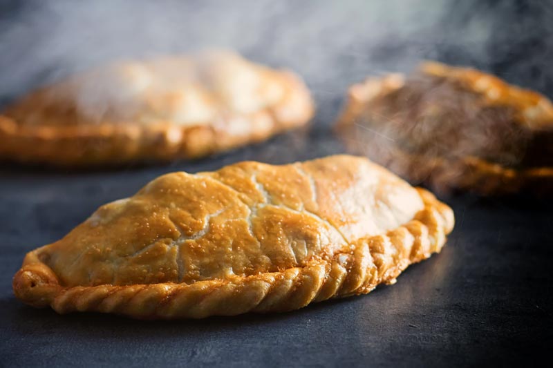 Landscape image of a traditional Cornish Pasty against a steamy dark back drop