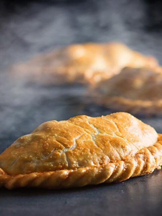 Square image of a traditional Cornish Pasty against a steamy dark back drop