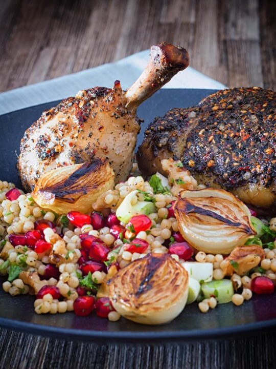 Portrait image of zaatar chicken legs served with giant couscous, pomegranate seeds and roasted shallots served on a black plate