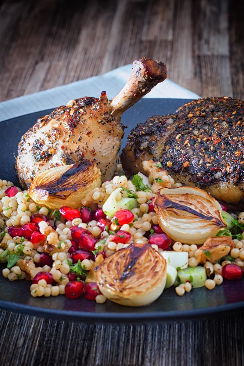 Portrait image of zaatar chicken legs served with giant couscous, pomegranate seeds and roasted shallots served on a black plate