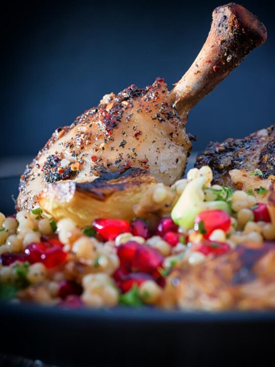 Close up portrait image of zaatar chicken legs served with giant couscous, pomegranate seeds