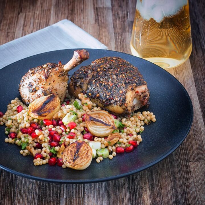 Square image of zaatar chicken legs served with giant couscous, pomegranate seeds and roasted shallots served on a black plate