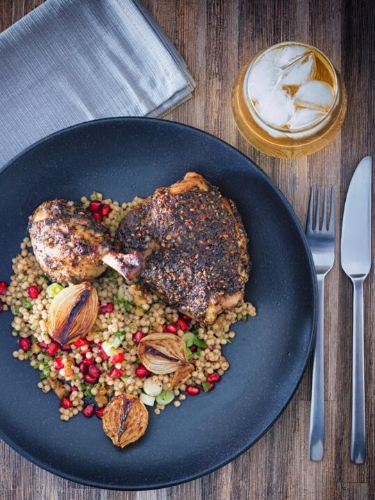 Portrait overhead image of zaatar chicken legs served with giant couscous, pomegranate seeds and roasted shallots served on a black plate