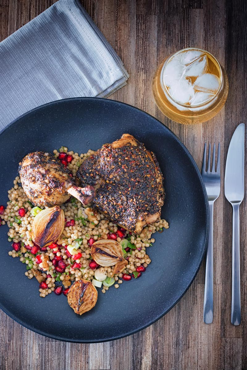Portrait overhead image of zaatar chicken legs served with giant couscous, pomegranate seeds and roasted shallots served on a black plate