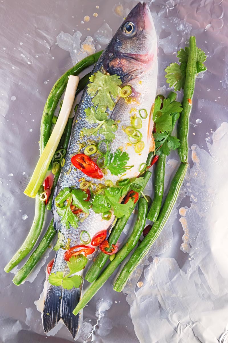 Portrait image of a raw whole sea bass with vegetables ready to be wrapped in a foil packet for fish en papillote