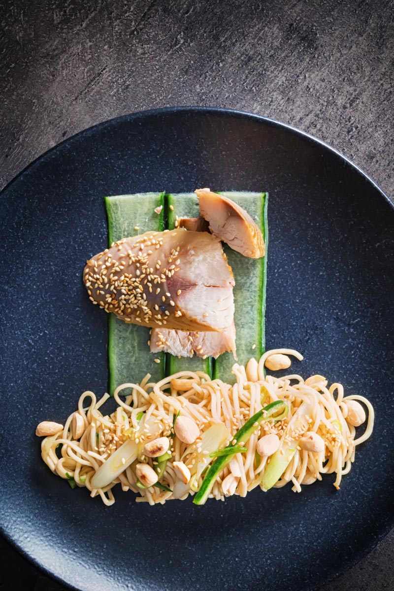 Portrait overhead image of a noodle salad featuring pickled cucumber and spring onions and sesame seeds served with smoked mackerel served on a black plate