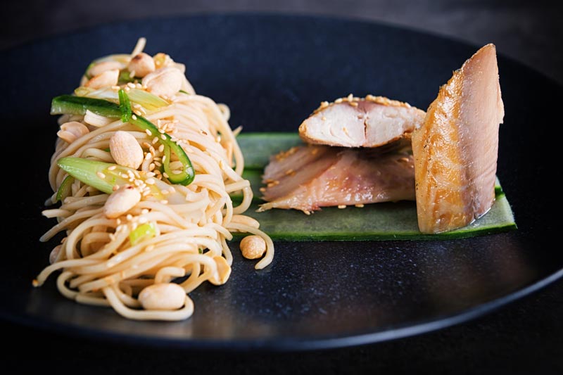 Landscape image of a noodle salad featuring pickled cucumber and spring onions and sesame seeds served with smoked mackerel served on a black plate