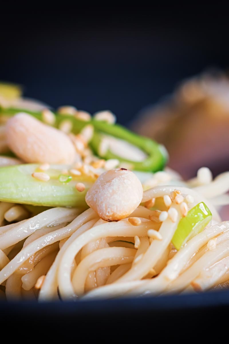 Close up portrait image of a noodle salad featuring pickled cucumber and spring onions and sesame seeds served on a black plate