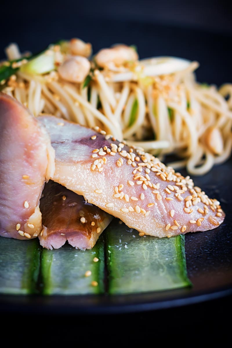Portrait image of a noodle salad featuring pickled cucumber and spring onions and sesame seeds served with smoked mackerel served on a black plate