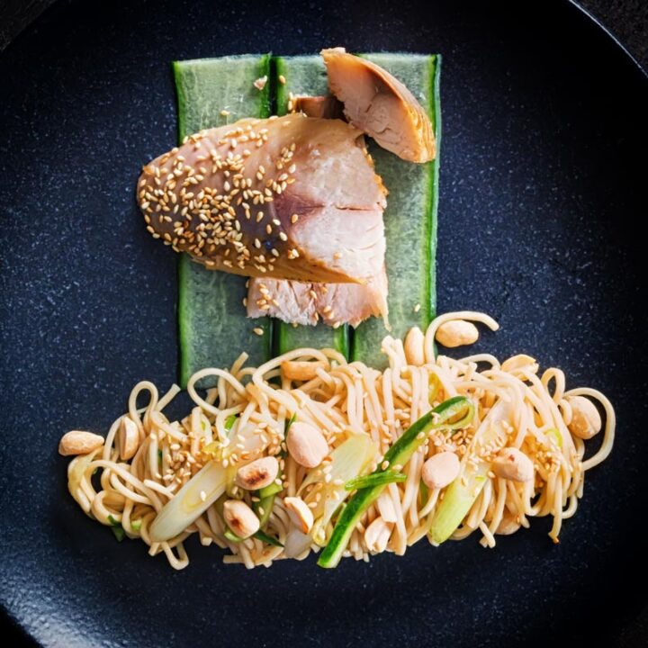 Square overhead image of a noodle salad featuring pickled cucumber and spring onions and sesame seeds served with smoked mackerel served on a black plate