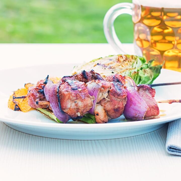 Square image of BBQ duck kebabs served on a white plate with grilled lettuce and seared orange segments served with a pint of beer