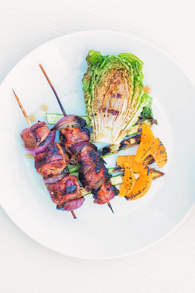 Portrait overhead image of BBQ duck kebabs served on a white plate with grilled lettuce, asparagus and seared orange segments