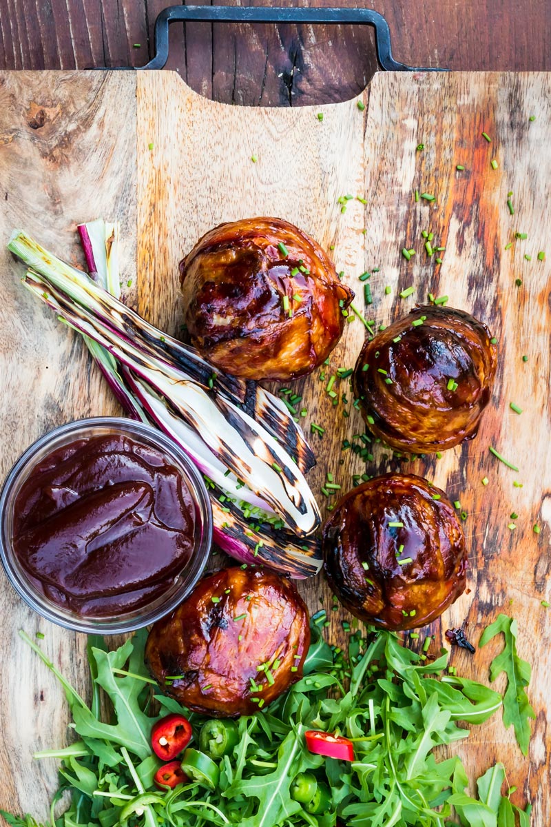 Portrait overhead image of Bacon wrapped Grilled Stuffed Onions served on a chopping board with BBQ sauce and a rocket salad