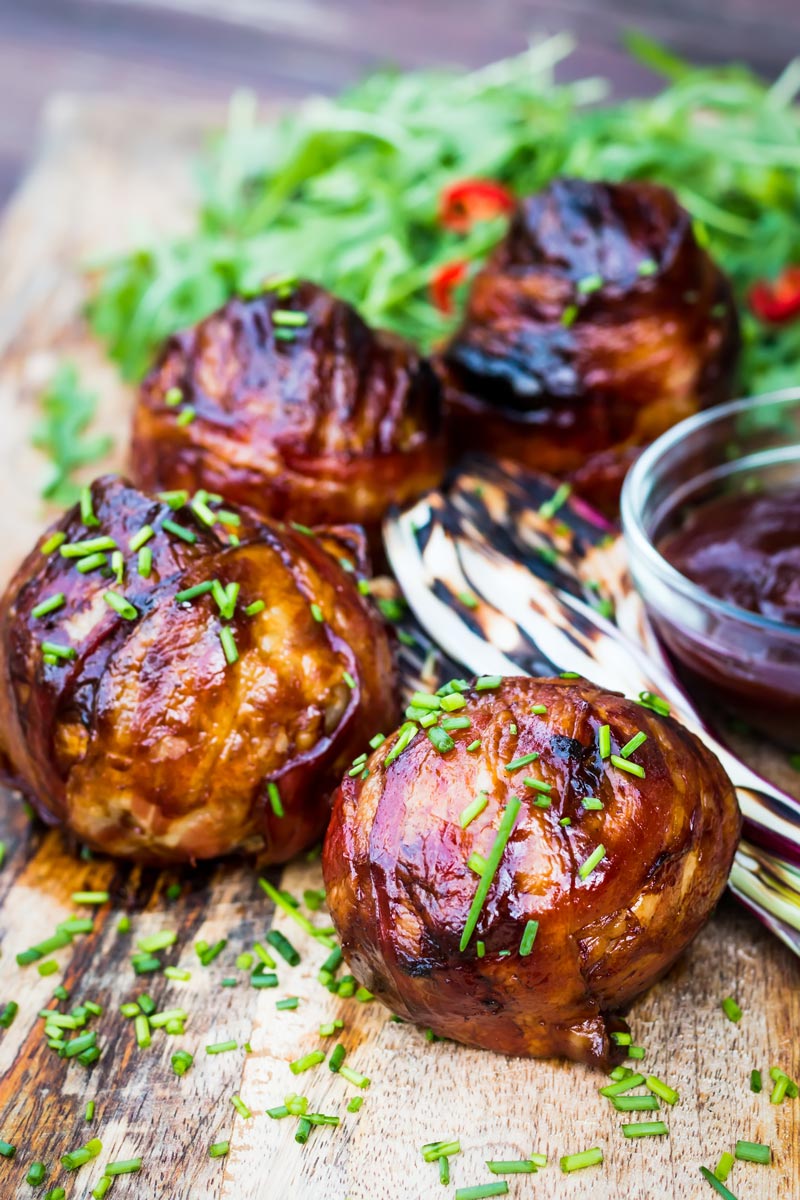 Portrait close up image of Bacon wrapped Grilled Stuffed Onions served on a chopping board with BBQ sauce and a rocket salad