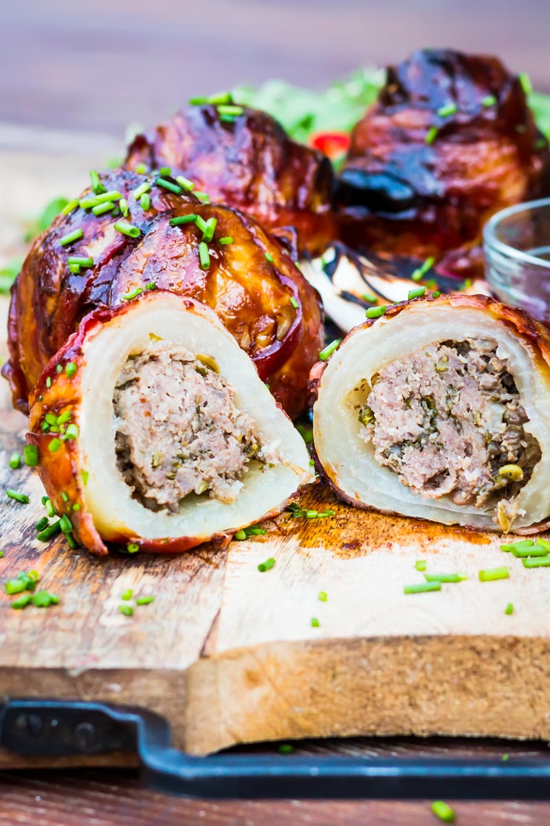 Portrait close up image of Bacon wrapped Grilled Stuffed Onions cut open and served on a chopping board with BBQ sauce and a rocket salad