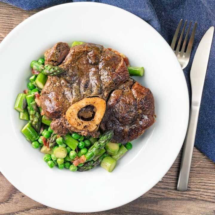 Square overhead image of slowly braised beef shin on the bone served on a bed of peas, asparagus and bacon served in a white bowl