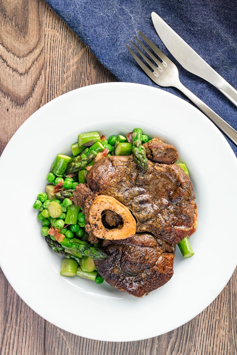 Portrait overhead image of slowly braised beef shin on the bone served on a bed of peas, asparagus and bacon served in a white bowl