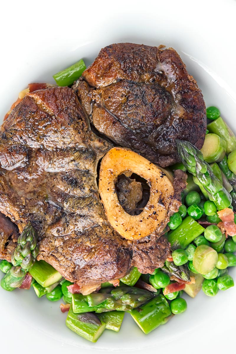 Portrait overhead close up image of slowly braised beef shin on the bone served on a bed of peas, asparagus and bacon served in a white bowl