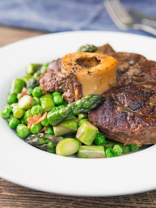Portrait image of slowly braised beef shin on the bone served on a bed of peas, asparagus and bacon served in a white bowl