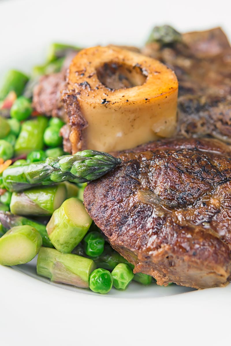 Portrait close up image of slowly braised beef shin on the bone served on a bed of peas, asparagus and bacon served in a white bowl