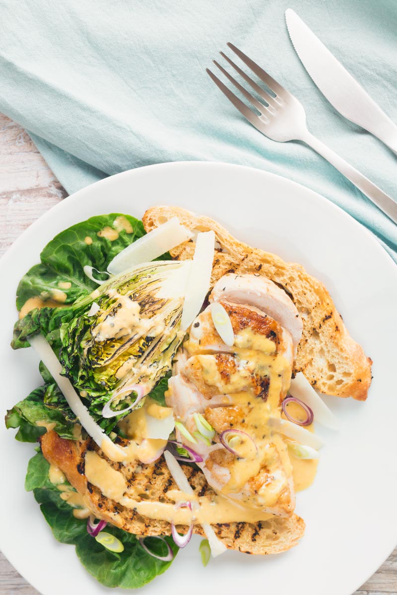 Portrait overhead image of a chicken Caesar Salad served on a white plate featuring a roast chicken breast, seared baby gem lettuce hearts, ciabatta croutons and parmesan shavings