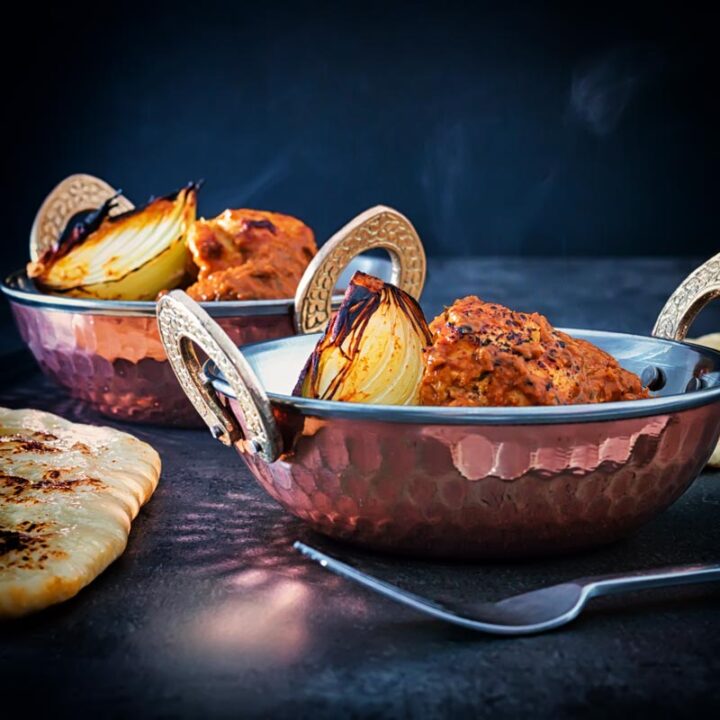 Square image of a chicken tikka masala curry served in two copper coated curry bowls served with naan bread against a dark backdrop
