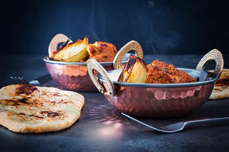 Landscape image of a chicken tikka masala curry served in two copper coated curry bowls served with naan bread against a dark backdrop