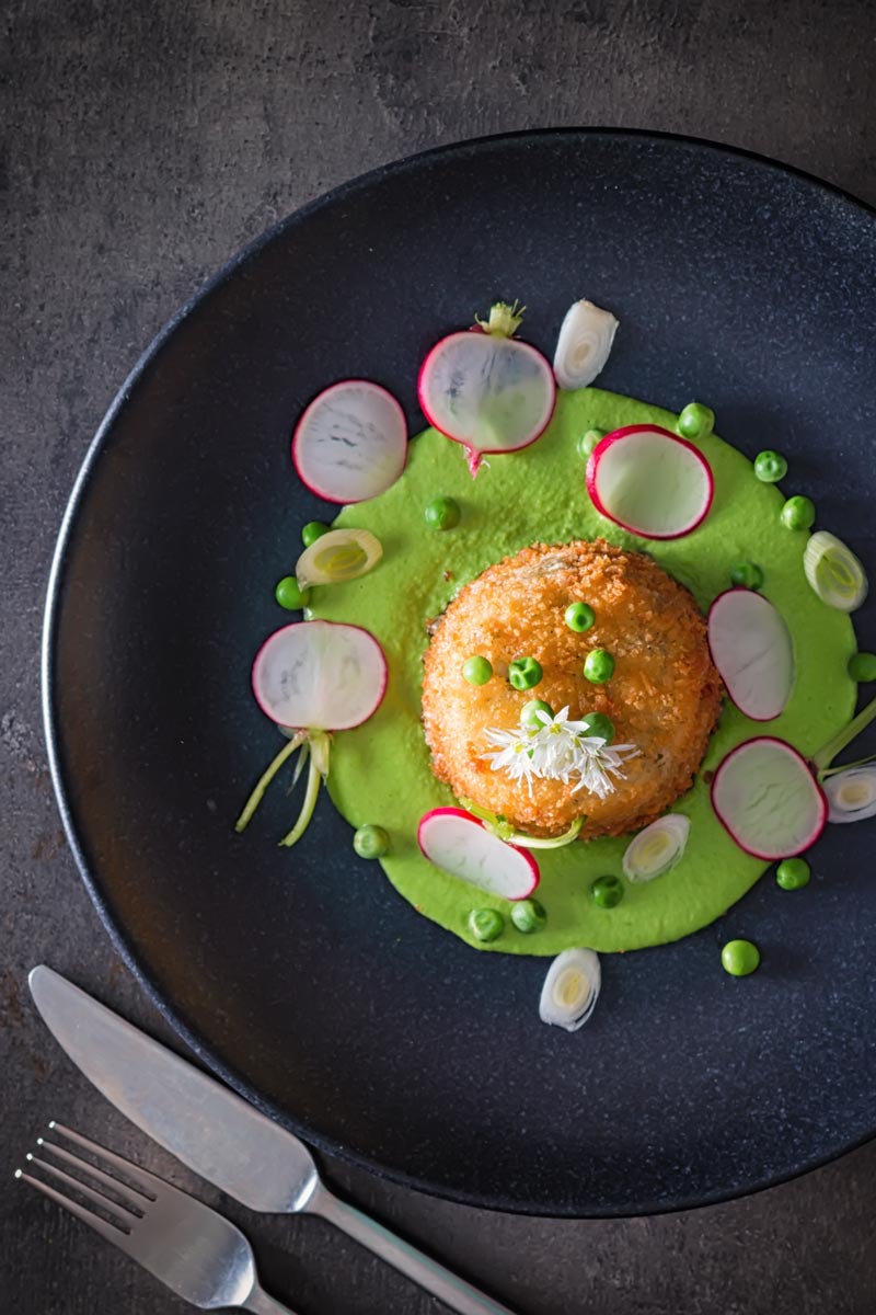 Portrait overhead image of golden deep fried goats cheese, served on pea puree with garlic flowers , sliced radish, peas and spring onions