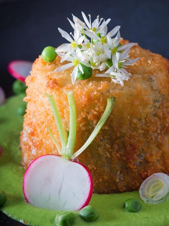 Portrait image of golden deep fried goats cheese, served on pea puree with garlic flowers , sliced radish, peas and spring onions
