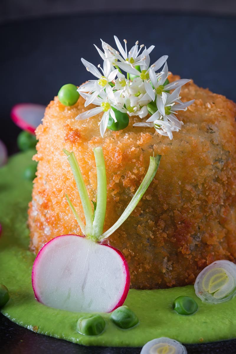Portrait image of golden deep fried goats cheese, served on pea puree with garlic flowers , sliced radish, peas and spring onions