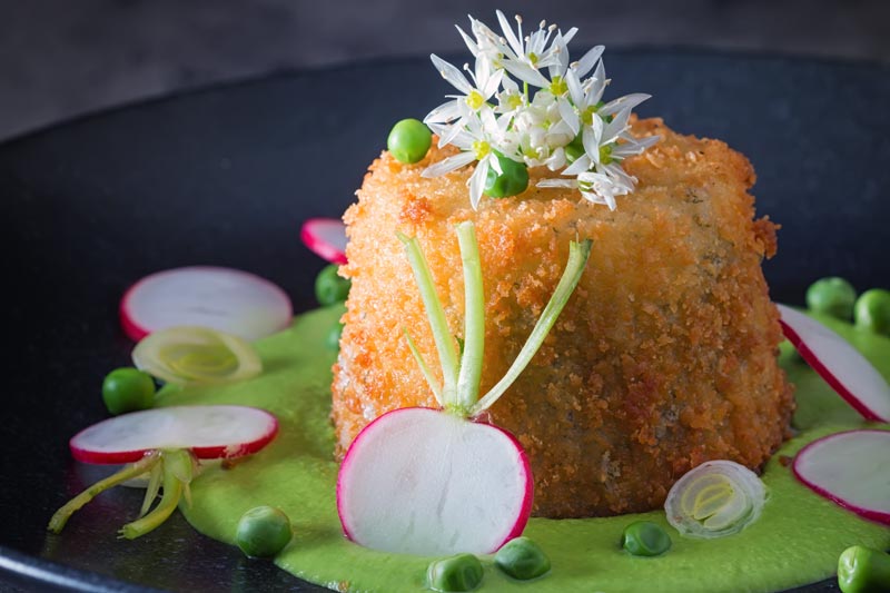 Landscape image of golden deep fried goats cheese, served on pea puree with garlic flowers , sliced radish, peas and spring onions
