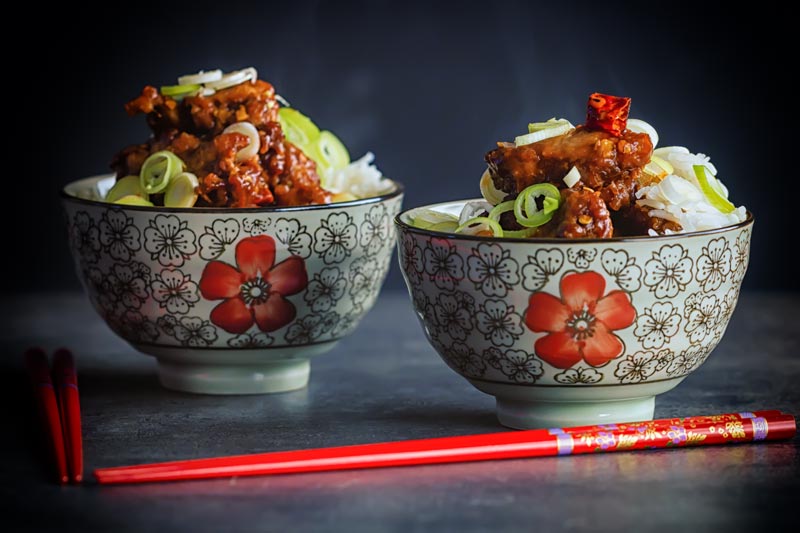 Landscape image of General Tso's chicken served in two small bowls decorated with a red flower with Asian styling and red chopsticks