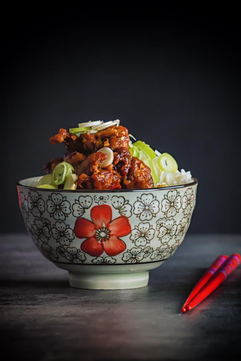 Portrait image of General Tso's chicken served in a bowl decorated with a red flower with Asian styling and red chopsticks