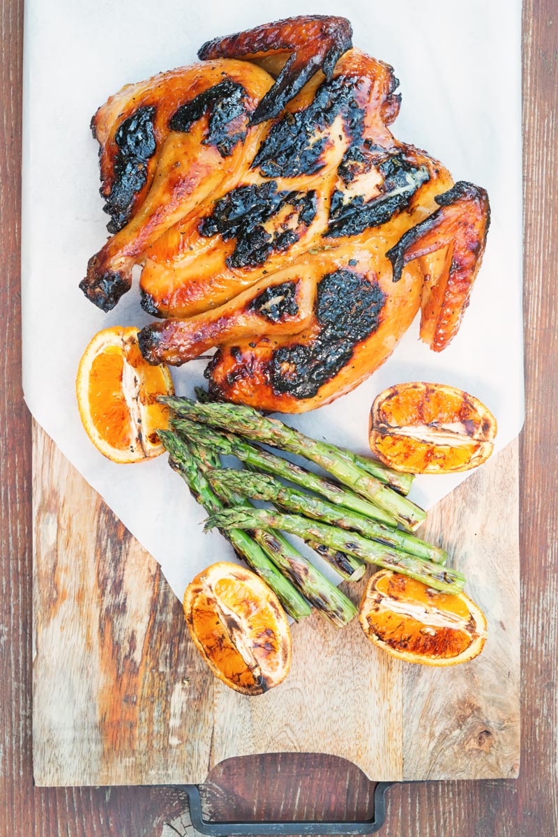 Portrait image of a Grilled Spatchcock Chicken served on a wooden chopping board with asparagus and grilled orange segments