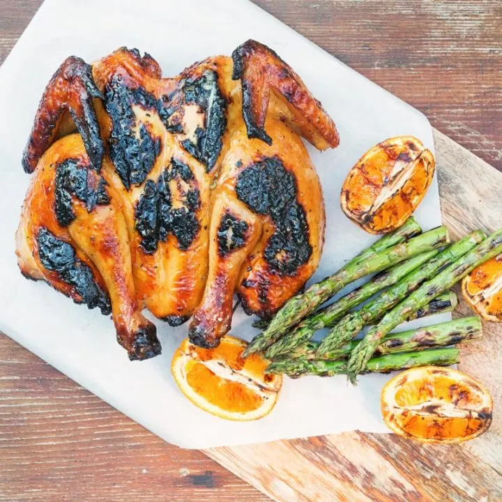 Square overhead image of a Grilled Spatchcock Chicken served on a wooden chopping board with asparagus and grilled orange segments