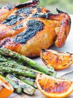 Portrait image of a Grilled Spatchcock Chicken served on a wooden chopping board with asparagus and grilled orange segments