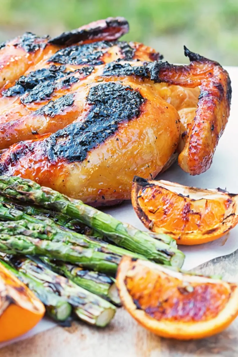 Portrait image of a Grilled Spatchcock Chicken served on a wooden chopping board with asparagus and grilled orange segments