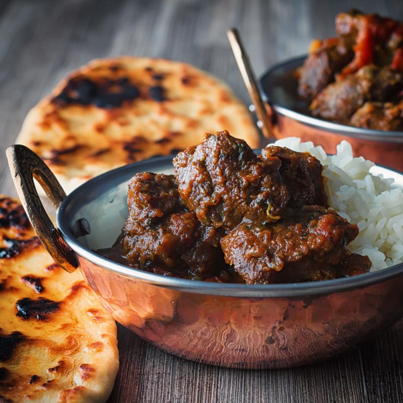 Square image of a beef vindaloo curry served in a copper coated curry bowl with a naan bread served on a rustic wooden table top