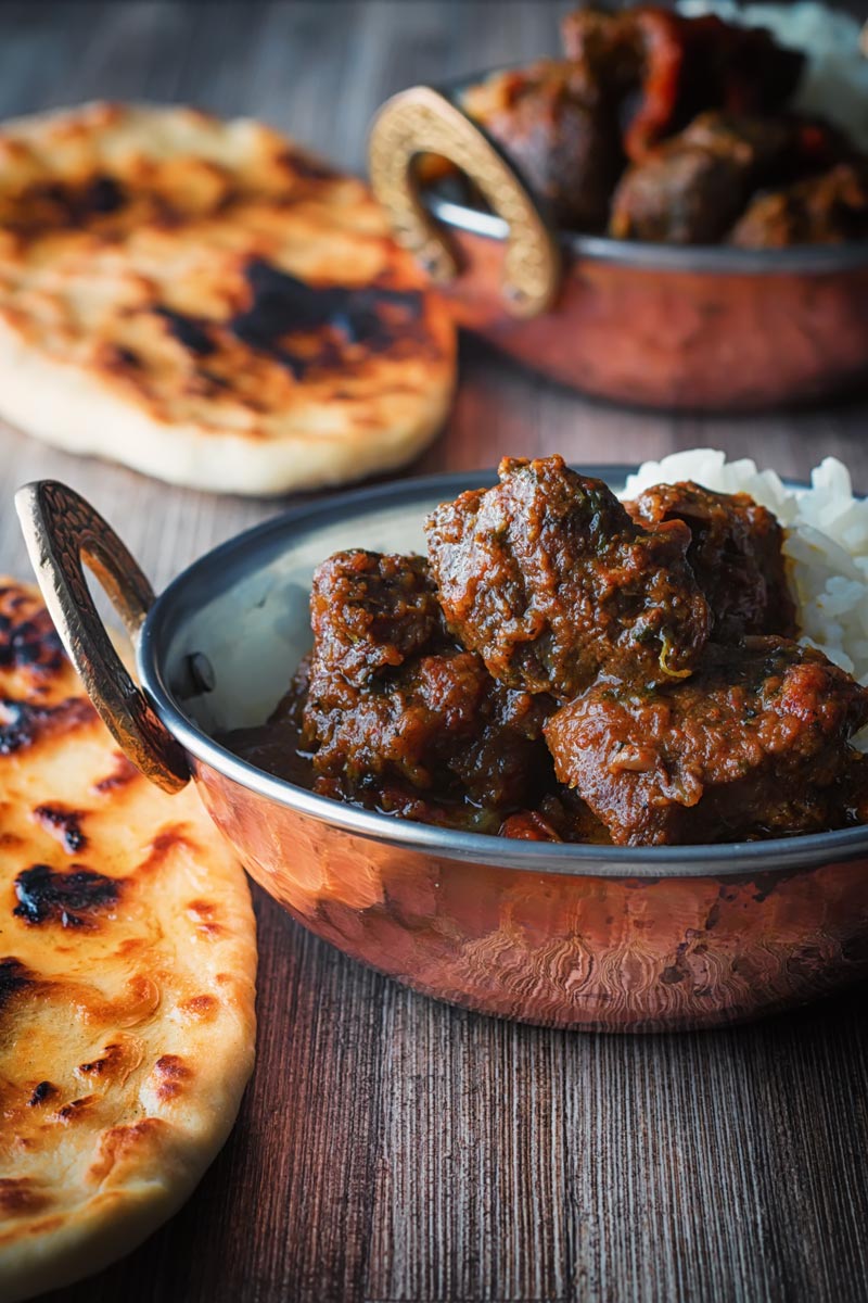 Portrait image of a beef vindaloo curry served in two copper coated curry bowls with a naan bread served on a rustic wooden table top