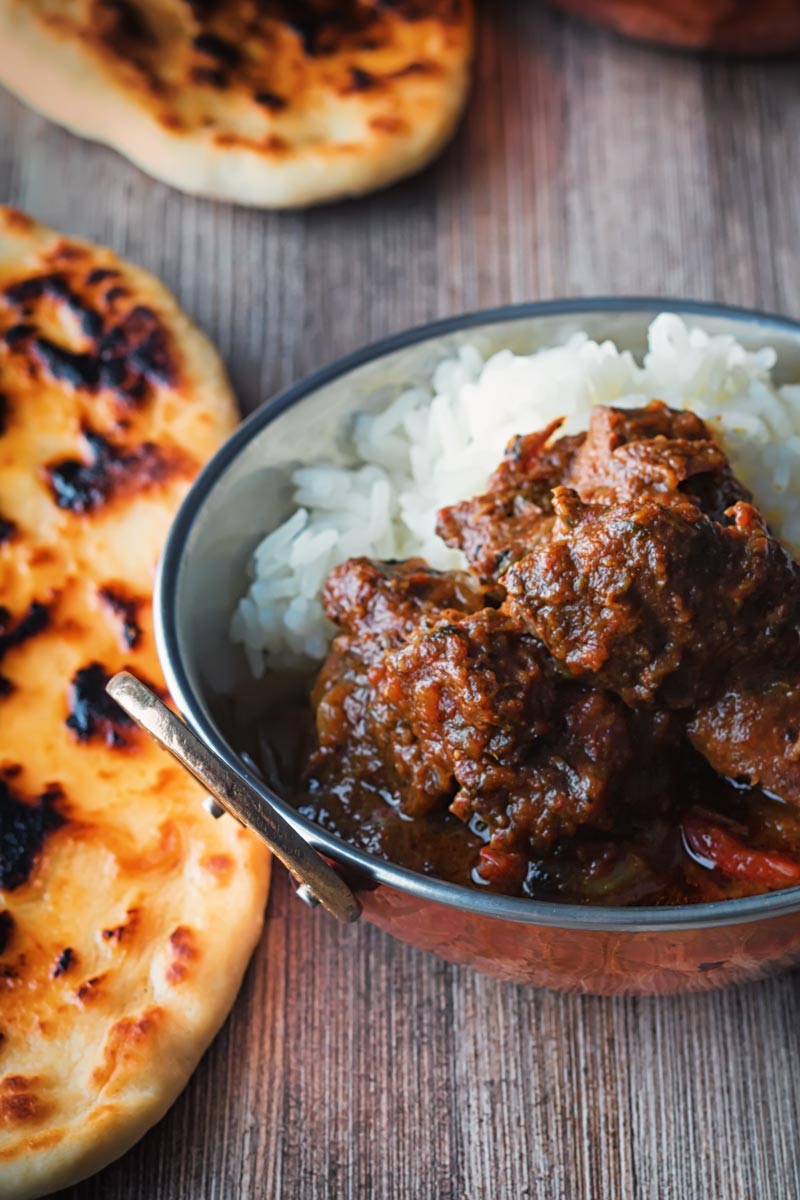 Portrait image of a beef vindaloo curry served in a copper coated curry bowls with a naan bread served on a rustic wooden table top taken from a high angle