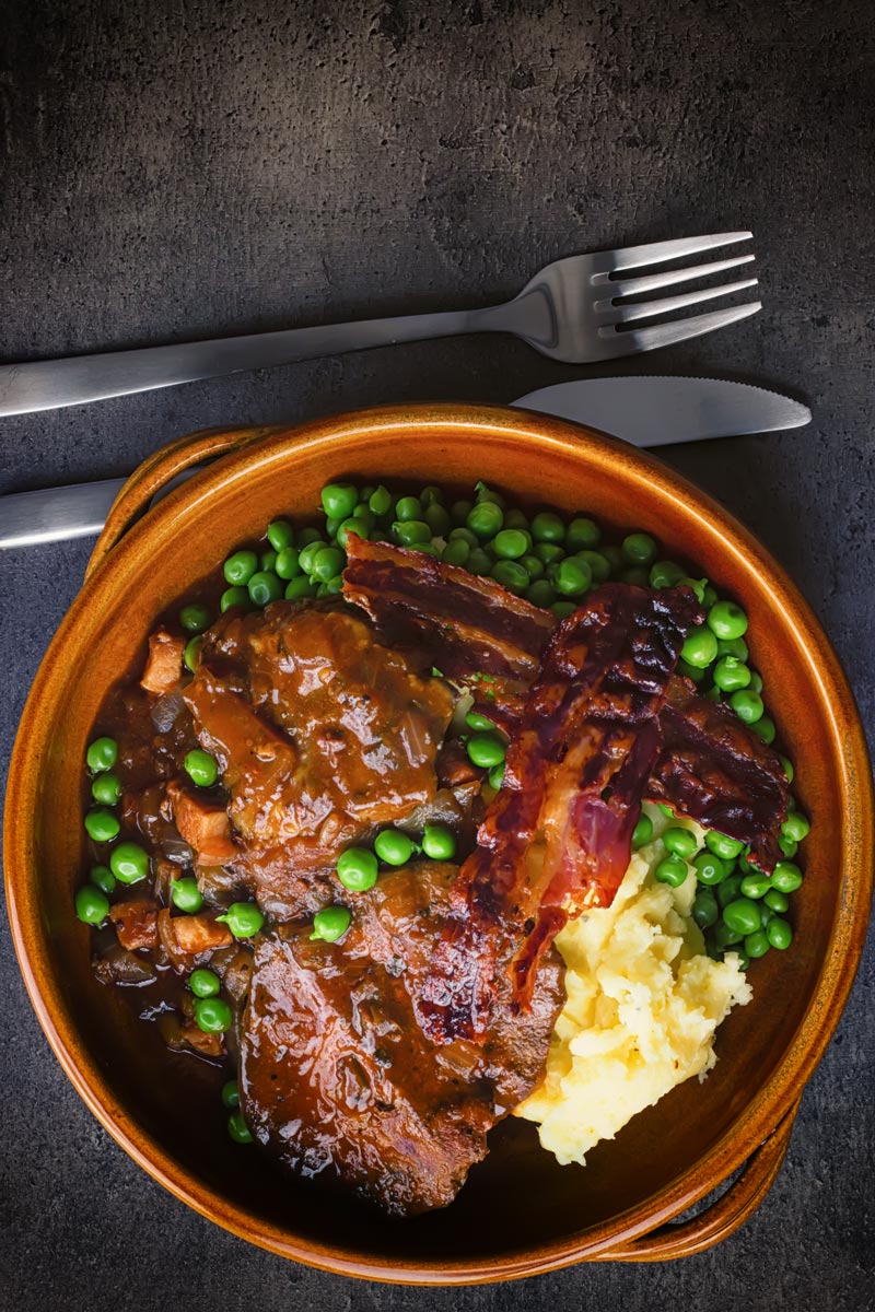 Portrait overhead image of liver and onions with crispy bacon, mashed potatoes and peas and onion gravy served in an earthenware bowl