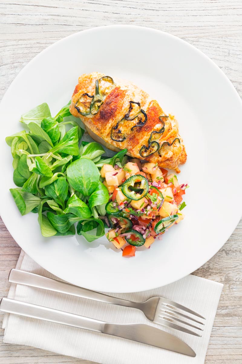 Portrait overhead image of a roasted jalapeño and cheddar cheese stuffed chicken breast served on a white plate with pineapple salsa and a green salad