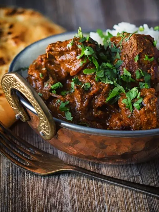 Portrait image of a lamb rogan josh curry served in a copper coated curry bowl with a naan bread
